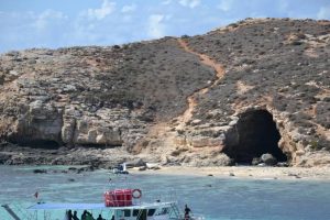 A foreground of very saturated blue sea with a small island in the back and a cave opening into it.