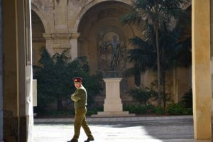 A guard walking on the courtyard of a palace, pillars and vegetation seen in the back