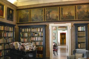 A 16th century green and brown accented library inside a decadent interior of a palace. A white hall into the rest of the palace is seen from a white doorway.