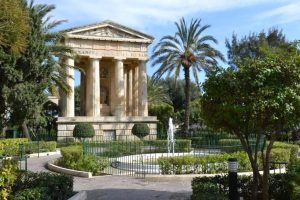 A garden with a pond and Greek reminiscent structure with pillars. Greenery all around, with palms framing the structure