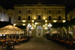 The Maltese National Library with a statue in front during nighttime. Scarcely lit and accompanied by two sets of outside cafe umbrellas on either side.