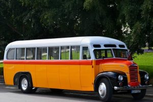Orange, red and white old bus in front of a green courtyard.