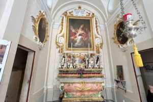 An upwards view of an altar, white walls in the chapel with decadent framing of a painting situated in the centre of the image