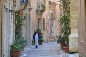 A curving street in Birgu with faint sunlight illuminating it, greenery spilling over the walls and a person walking down the street carrying bags