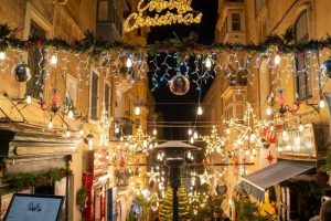 A crowded Valletta street with lights and banners hanging above head level and a "Merry Christmas" sign in lights in the center of the image