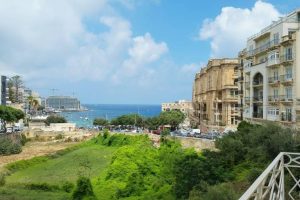A perspective image of Balluta Bay from a far away viewpoint with greenery and a fraction of the city skyline visible.